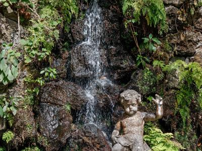 A stone statue of a seated cherub or child figure positioned in front of a small, cascading waterfall surrounded by lush ferns and tropical foliage.
