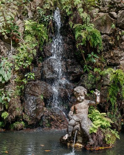 A stone statue of a seated cherub or child figure positioned in front of a small, cascading waterfall surrounded by lush ferns and tropical foliage.