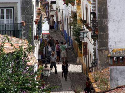 A high-angle view looking down a narrow, cobblestone pedestrian street lined with whitewashed buildings featuring terracotta tile roofs, flower boxes, and stone accents, with several tourists walking along the path.