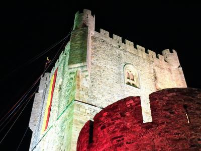 A low-angle night shot of a medieval stone castle tower illuminated with green and red lights, featuring a Spanish flag hanging from the battlements.