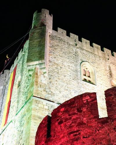 A low-angle night shot of a medieval stone castle tower illuminated with green and red lights, featuring a Spanish flag hanging from the battlements.