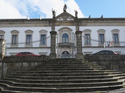 A large white neoclassical building with a central pediment featuring statues, a stone staircase leading up to a gated entrance, and flags flying on the right side.