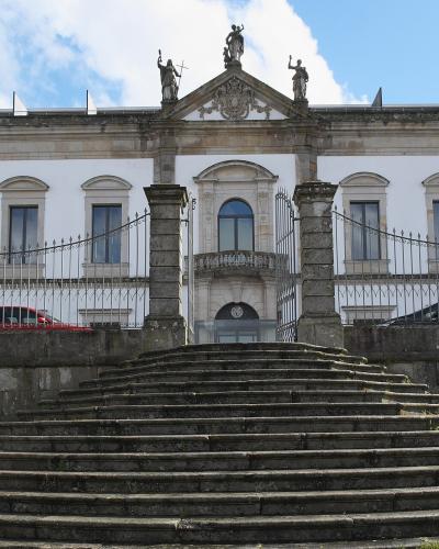 A large white neoclassical building with a central pediment featuring statues, a stone staircase leading up to a gated entrance, and flags flying on the right side.