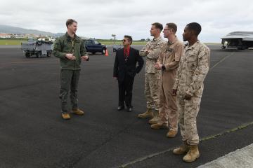 The image shows a group of military personnel and a civilian in a suit standing on an airfield tarmac, with a B-2 Spirit stealth bomber visible in the background and a pilot in flight gear speaking to them.