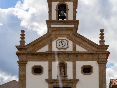 A low-angle photograph of a white church facade with stone trim, a bell tower, a clock, and a statue niche, set against a cloudy blue sky.