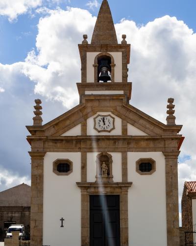 A low-angle photograph of a white church facade with stone trim, a bell tower, a clock, and a statue niche, set against a cloudy blue sky.