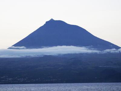 A large, conical stratovolcano rising above a band of low clouds, viewed from across a body of water with a coastal settlement visible at the base.
