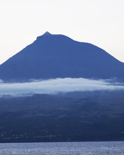A large, conical stratovolcano rising above a band of low clouds, viewed from across a body of water with a coastal settlement visible at the base.
