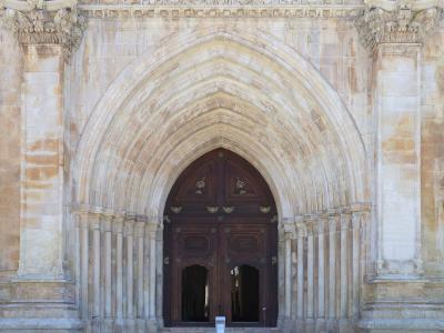 A close-up view of a large, ornate stone portal featuring a pointed Gothic arch with multiple recessed moldings (archivolts), flanked by columns with carved capitals and topped by a decorative frieze, leading to a dark wooden double door.