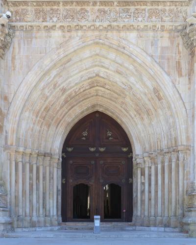 A close-up view of a large, ornate stone portal featuring a pointed Gothic arch with multiple recessed moldings (archivolts), flanked by columns with carved capitals and topped by a decorative frieze, leading to a dark wooden double door.