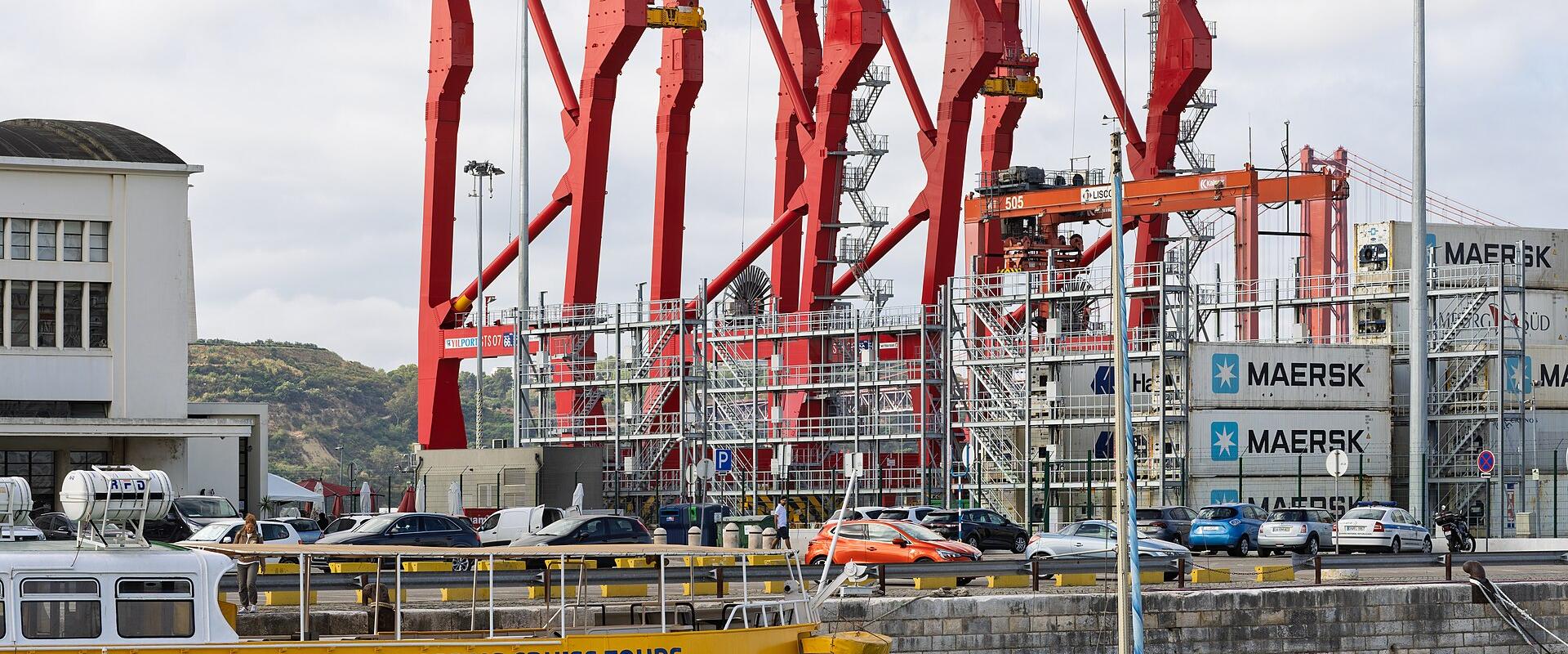 A yellow sightseeing cruise boat and a white sailboat are in the foreground of a harbor, with large red port cranes and shipping containers in the background.