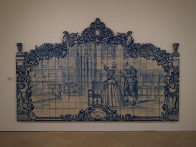 A large, ornate blue-and-white azulejo tile panel depicting a historical scene with figures in period dress, framed by elaborate Baroque-style tilework, mounted on a plain white wall inside a museum gallery.