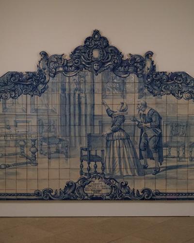 A large, ornate blue-and-white azulejo tile panel depicting a historical scene with figures in period dress, framed by elaborate Baroque-style tilework, mounted on a plain white wall inside a museum gallery.