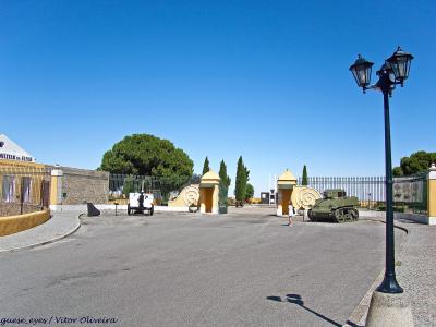 A paved entrance area with a yellow gatehouse and fence, featuring a military tank on display and a sign on the left building.
