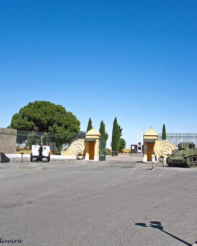 A paved entrance area with a yellow gatehouse and fence, featuring a military tank on display and a sign on the left building.