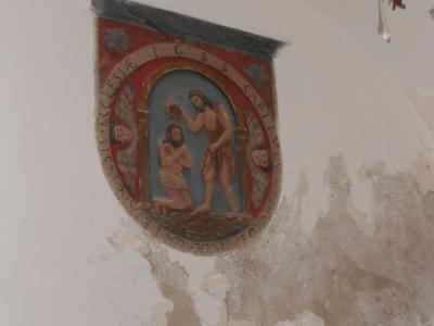 A close-up interior shot of a painted religious relief or mural on a white plaster wall, depicting the Baptism of Christ with Latin inscriptions including "IGS" and "CAPITULUM," alongside a glass chandelier and peeling paint.