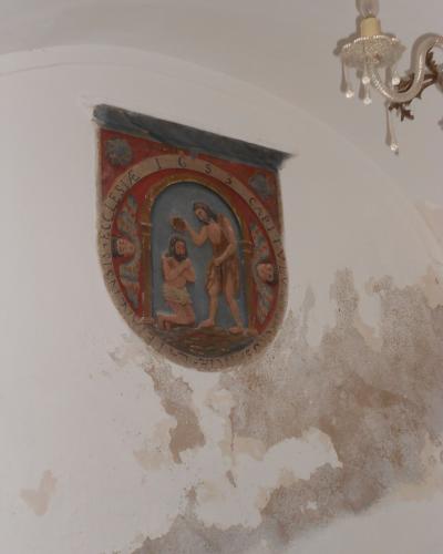 A close-up interior shot of a painted religious relief or mural on a white plaster wall, depicting the Baptism of Christ with Latin inscriptions including "IGS" and "CAPITULUM," alongside a glass chandelier and peeling paint.