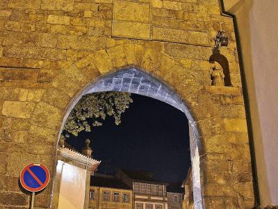 A night photograph of a stone archway gate with a coat of arms above the arch, looking out onto a lit street with traditional buildings and a cobblestone road.