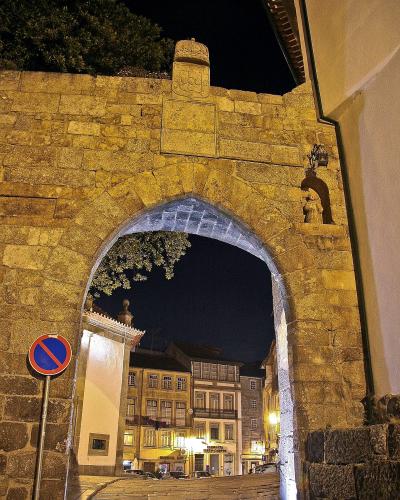 A night photograph of a stone archway gate with a coat of arms above the arch, looking out onto a lit street with traditional buildings and a cobblestone road.