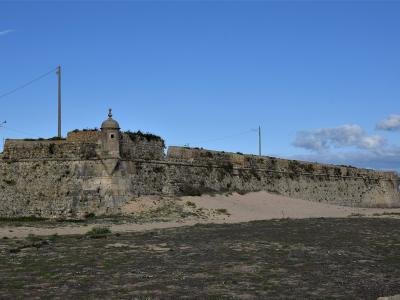 A low-angle view of a historic stone fortress wall with a central sentry box (garita) and battlements, situated on a sandy beach next to the ocean under a blue sky.
