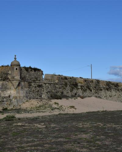 A low-angle view of a historic stone fortress wall with a central sentry box (garita) and battlements, situated on a sandy beach next to the ocean under a blue sky.