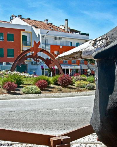 A street scene in a Portuguese town featuring a roundabout with decorative metal arches, colorful buildings, and a large dark sculpture in the foreground.
