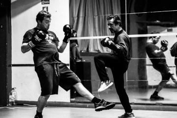 A black and white street photograph showing two men sparring in a boxing ring, with one man kicking and the other blocking; signage reading "SAÍDA" (Portuguese for "Exit") is visible on the wall.