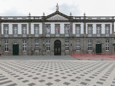 A large, two-story neoclassical building with a stone facade, white upper walls, and a central pediment featuring a statue, situated behind a plaza with distinctive black-and-white wave-patterned pavement.