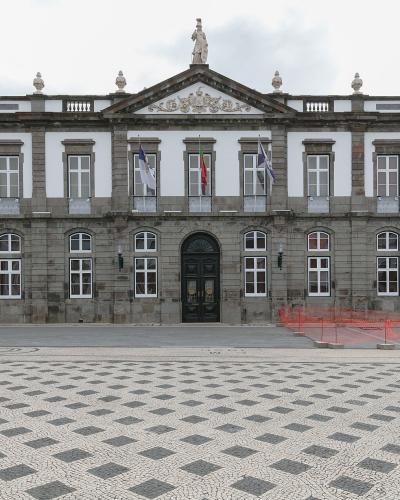 A large, two-story neoclassical building with a stone facade, white upper walls, and a central pediment featuring a statue, situated behind a plaza with distinctive black-and-white wave-patterned pavement.