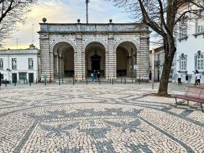 A stone building with three large arches (a porch or loggia) situated in a paved town square featuring a large cobblestone mosaic of the Portuguese coat of arms.