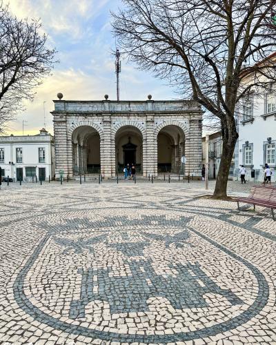 A stone building with three large arches (a porch or loggia) situated in a paved town square featuring a large cobblestone mosaic of the Portuguese coat of arms.