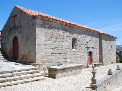 A stone church with a red-tiled roof, a large arched wooden door on the left, and a smaller door on the right, situated on a cobblestone plaza with stone benches and steps.
