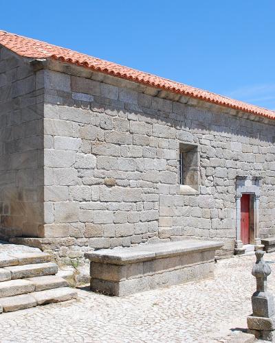 A stone church with a red-tiled roof, a large arched wooden door on the left, and a smaller door on the right, situated on a cobblestone plaza with stone benches and steps.