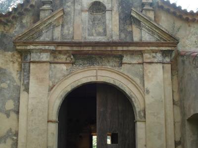 A weathered, historic stone facade featuring a large arched wooden doorway and a decorative upper section with a niche and circular window, set against a bright sky with tree branches visible.