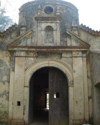 A weathered, historic stone facade featuring a large arched wooden doorway and a decorative upper section with a niche and circular window, set against a bright sky with tree branches visible.
