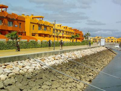 A waterfront promenade lined with distinctive bright orange and yellow apartment buildings, featuring a rocky embankment and calm water channel.
