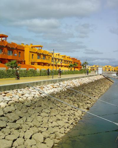 A waterfront promenade lined with distinctive bright orange and yellow apartment buildings, featuring a rocky embankment and calm water channel.