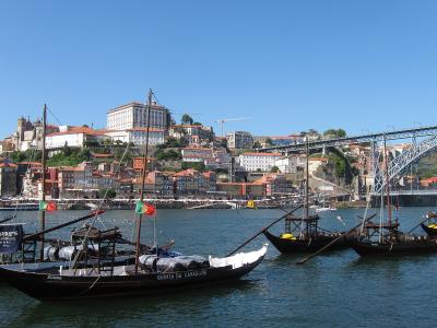 A view of the Douro River waterfront in Porto, Portugal, featuring several traditional wooden Rabelo boats moored in the foreground with the iconic Dom Luís I Bridge and the Ribeira district hillside in the background.