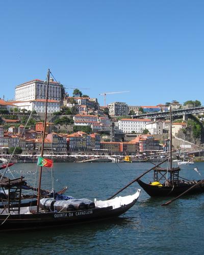 A view of the Douro River waterfront in Porto, Portugal, featuring several traditional wooden Rabelo boats moored in the foreground with the iconic Dom Luís I Bridge and the Ribeira district hillside in the background.