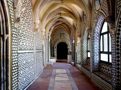 A long, vaulted corridor with Gothic-style ribbed ceilings and walls covered in intricate black, white, and blue geometric azulejo tilework, featuring arched windows on the right and a dark doorway at the far end.
