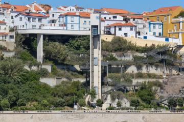 A harbor scene in Sines, Portugal, showing fishing boats in the foreground, a modern elevator structure connecting the lower port area to the upper town, and white buildings with terracotta roofs on a hillside under a clear blue sky.