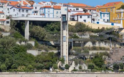 A harbor scene in Sines, Portugal, showing fishing boats in the foreground, a modern elevator structure connecting the lower port area to the upper town, and white buildings with terracotta roofs on a hillside under a clear blue sky.