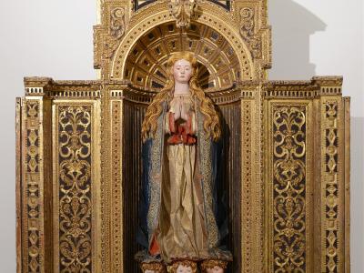 A gilded wooden altarpiece featuring a central statue of the Virgin Mary with long flowing hair, set within an ornate architectural frame with painted panels at the base, displayed against a plain white wall.