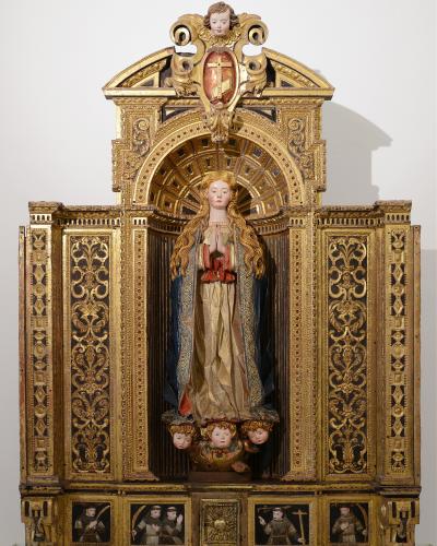 A gilded wooden altarpiece featuring a central statue of the Virgin Mary with long flowing hair, set within an ornate architectural frame with painted panels at the base, displayed against a plain white wall.